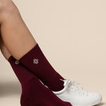 Burgundy socks with a logo worn by a person with a white sneaker on a beige background