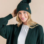Modal wearing chunky knit Adult pom beanie in cypress and chunky knit oversized cardigan in cypress over white t-shirt on a beige background