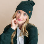 Modal wearing chunky knit Adult pom beanie in cypress and chunky knit oversized cardigan in cypress over white t-shirt on a beige background
