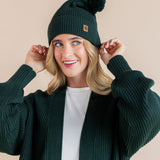 Modal posing in chunky knit Adult pom beanie in cypress and chunky knit oversized cardigan in cypress over white t-shirt on a beige background