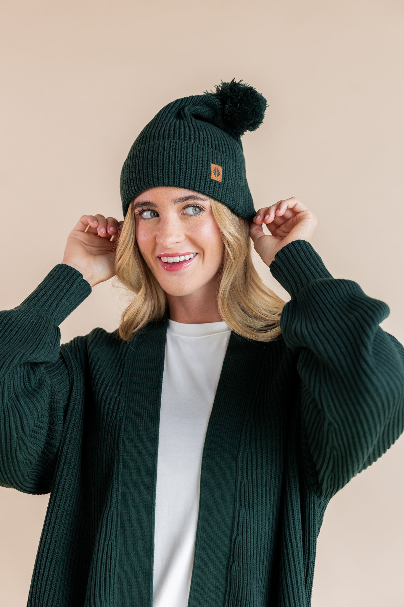 Modal posing in chunky knit Adult pom beanie in cypress and chunky knit oversized cardigan in cypress over white t-shirt on a beige background