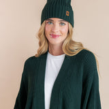 Modal wearing chunky knit Adult ribbed beanie in cypress and chunky knit oversized cardigan in cypress over white t-shirt on a beige background