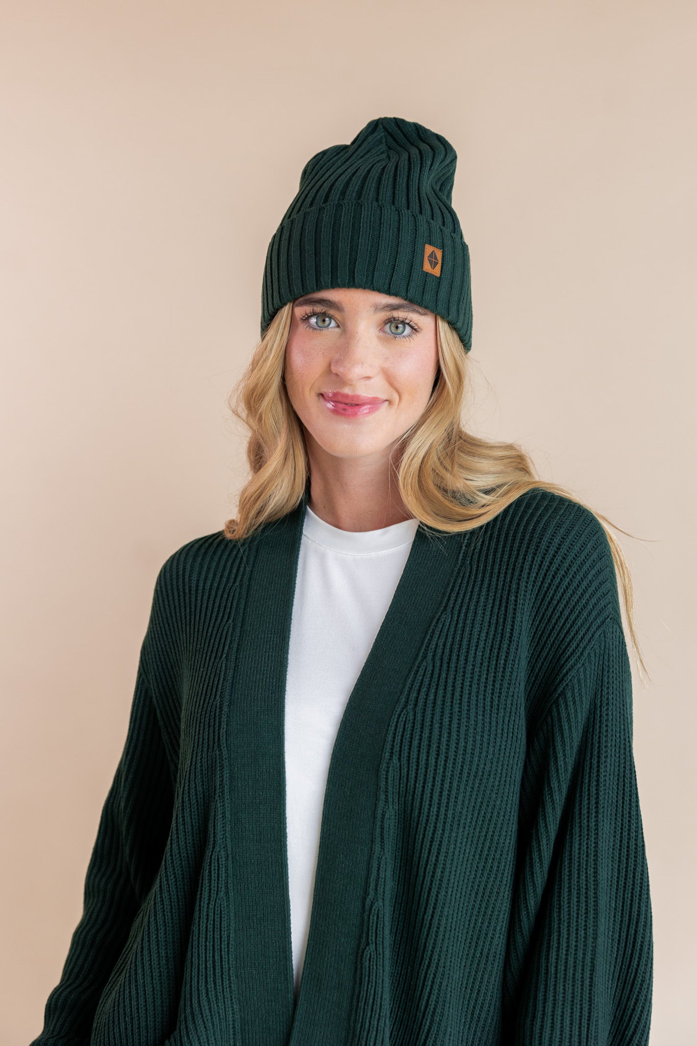 Modal wearing chunky knit Adult ribbed beanie in cypress and chunky knit oversized cardigan in cypress over white t-shirt on a beige background