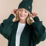 Modal wearing chunky knit Adult ribbed beanie in cypress and chunky knit oversized cardigan in cypress over white t-shirt on a beige background