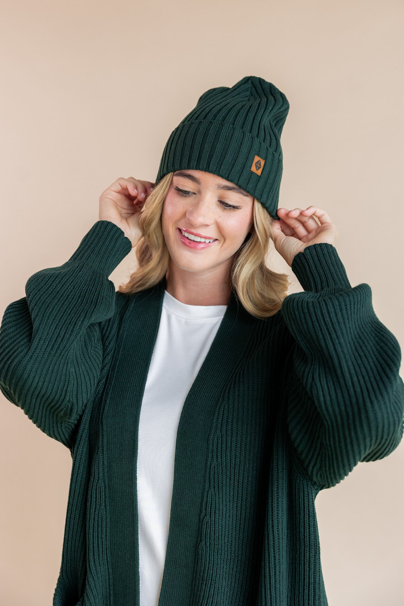 Modal wearing chunky knit Adult ribbed beanie in cypress and chunky knit oversized cardigan in cypress over white t-shirt on a beige background