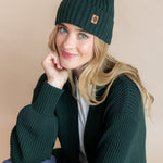 Modal wearing chunky knit Adult ribbed beanie in cypress over chunky knit oversized cardigan in cypress on a beige background