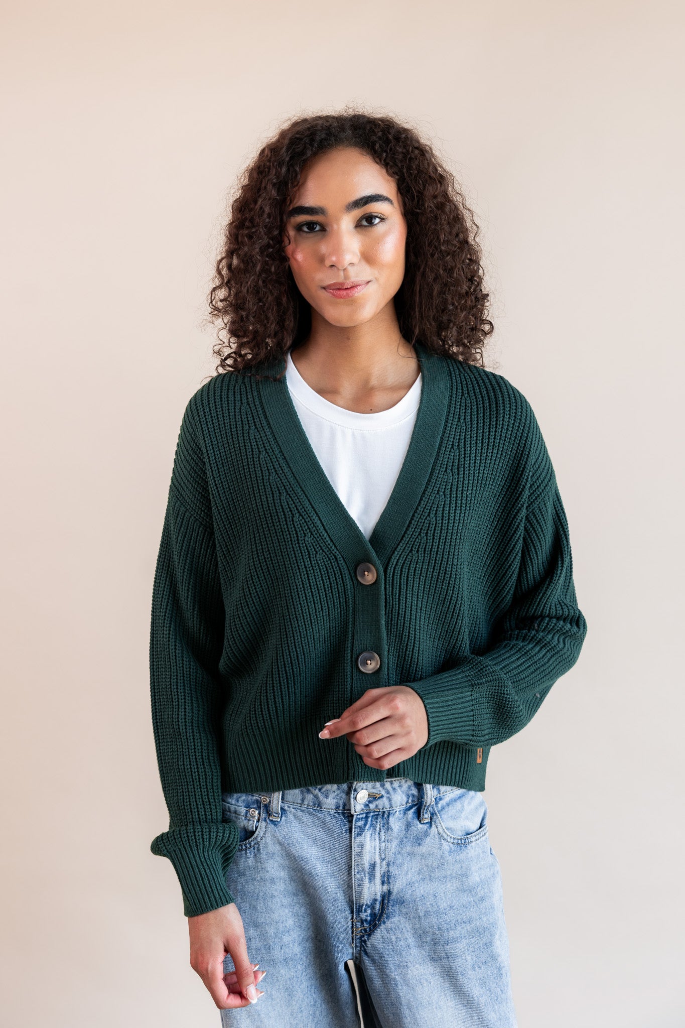 Modal posing in Chunky Knit Cropped Cardigan in cypress over white t-shirt and blue jeans on a beige background