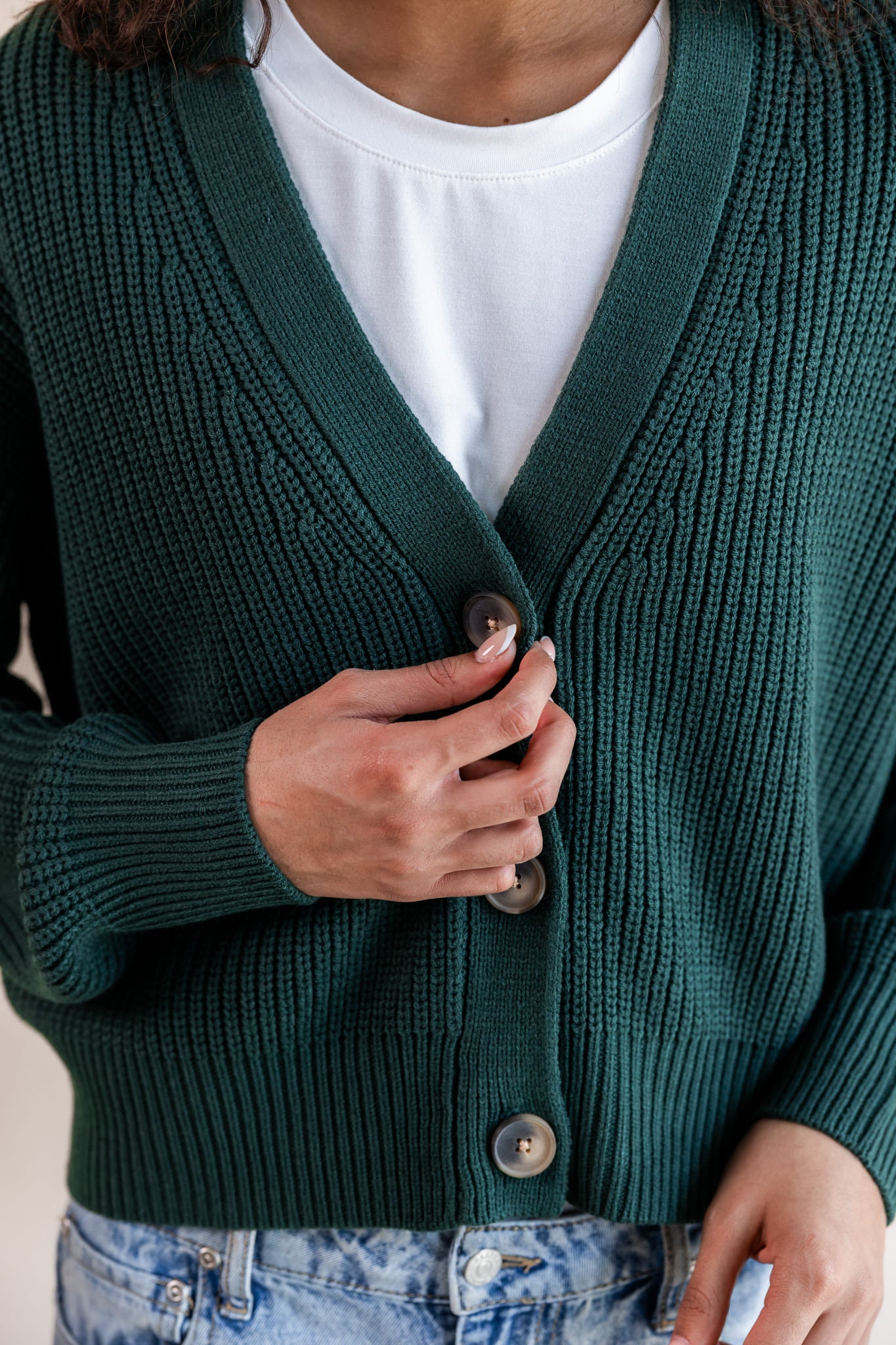 Modal holding the button of Chunky Knit Cropped Cardigan in cypress over white t-shirt and blue jeans on a beige background