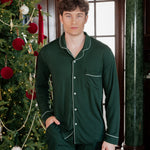 A man wearing a dark green long-sleeve pajama set with white piping stands in a decorated indoor setting next to a Christmas tree.