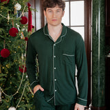 A man wearing a dark green long-sleeve pajama set with white piping stands in a decorated indoor setting next to a Christmas tree.