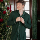 A man wearing a dark green long-sleeve pajama set with white piping stands in front of a window and a decorated Christmas tree.