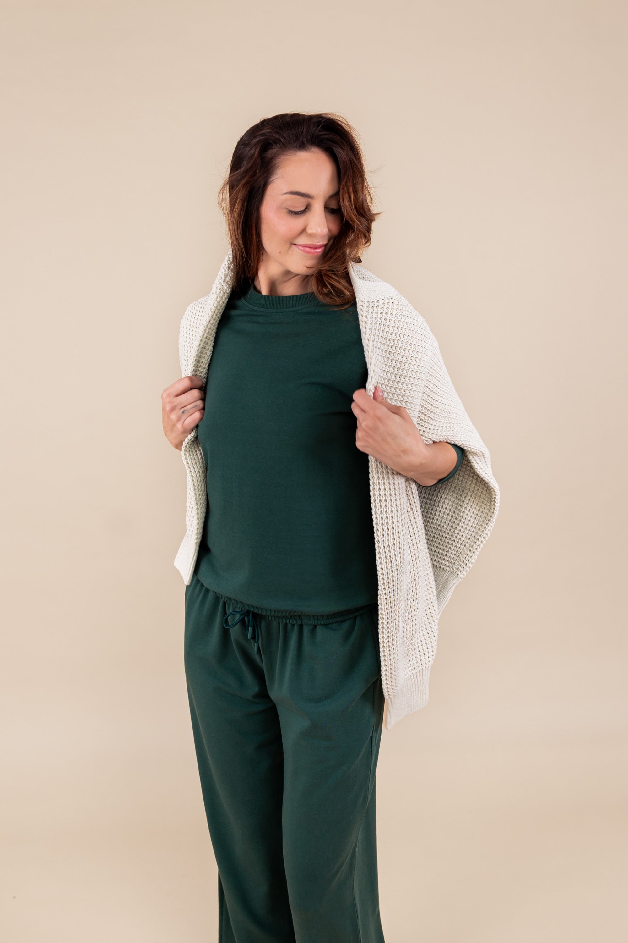 Woman wearing Women's Sweatpants in Cypress color under similar color women's sweatshirt and a beige sweater over her shoulders on a beige background