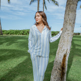 Woman in light blue striped pajama set standing next to a palm tree on a grassy area.