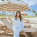 Woman in a light blue outfit sitting on a beach chair under a striped umbrella with palm trees and a clear sky in the background.