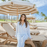 Woman in a light blue outfit sitting on a beach chair under a striped umbrella with palm trees and a clear sky in the background.