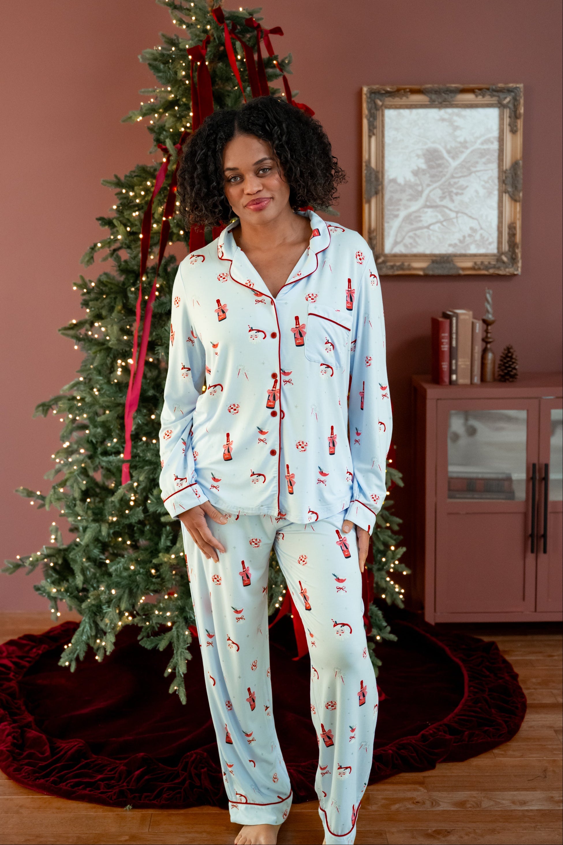 Woman wearing a blue pajama set with a festive print of santas, champagne, mugs, and disco balls standing in front of a christmas tree. 
---1605 3