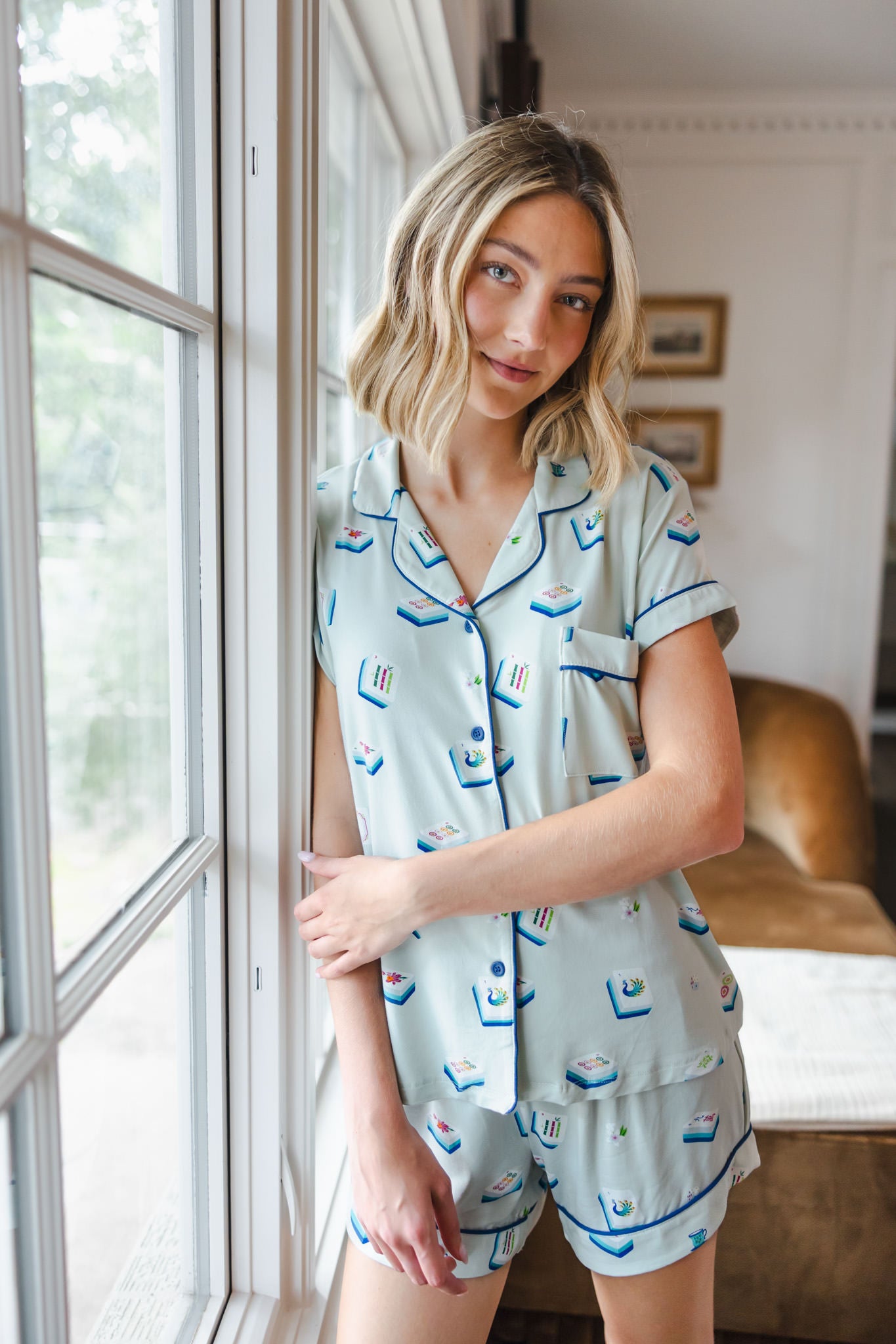 woman wearing light green short sleeve pajama set with mahjong tile print standing against a window

