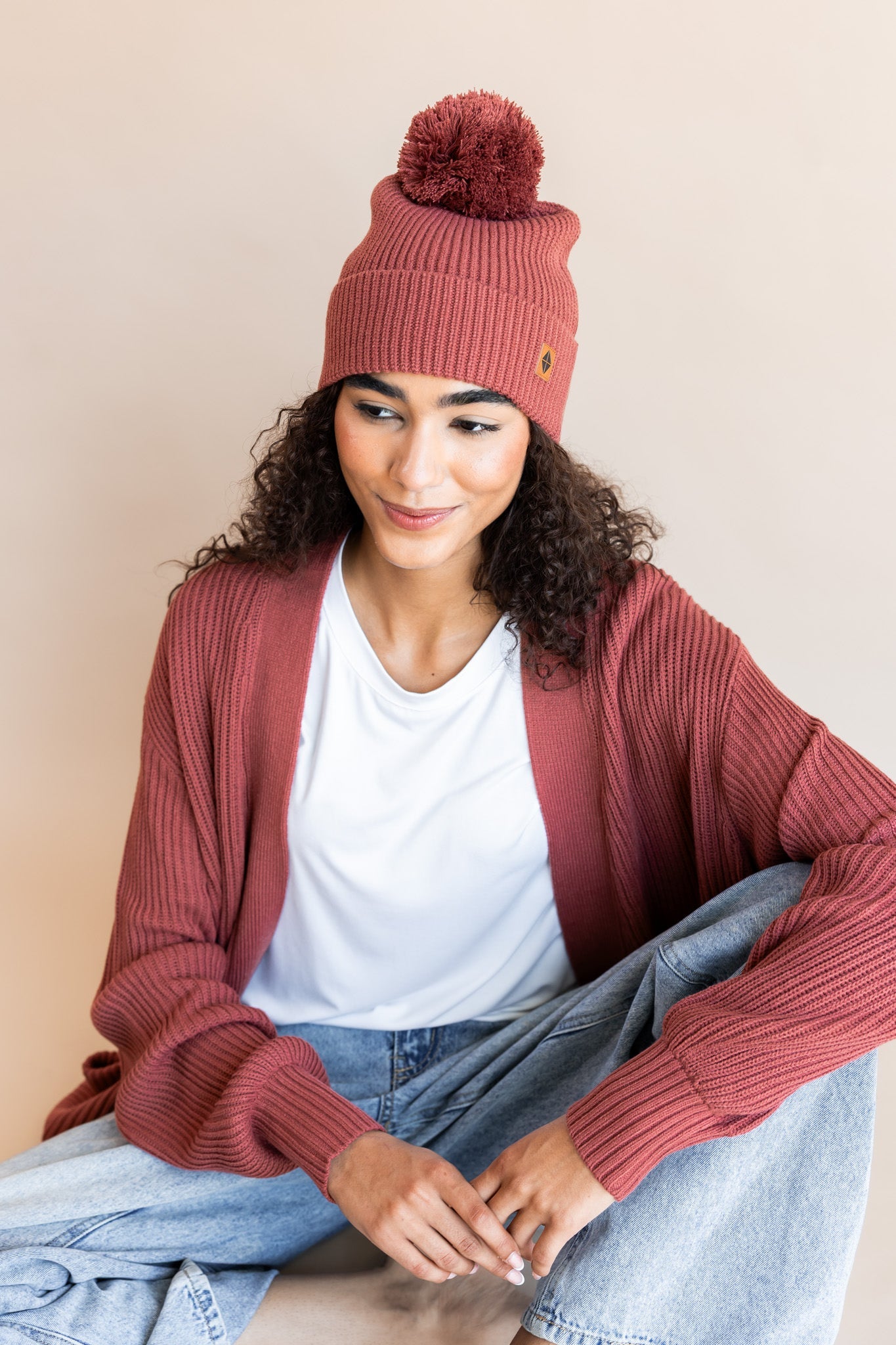 Modal wearing Chunky Knit Adult Pom in ember over white t-shirt and chunky knit women's cardigan in ember and blue jeans on a beige background