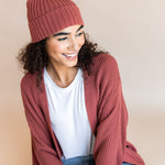 Modal wearing Chunky Knit Adult Ribbed Beanie in ember over white t-shirt and chunky knit women's cardigan in ember and blue jeans on a beige background