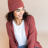 Modal wearing Chunky Knit Adult Ribbed Beanie in ember over white t-shirt and chunky knit women's cardigan in ember and blue jeans on a beige background