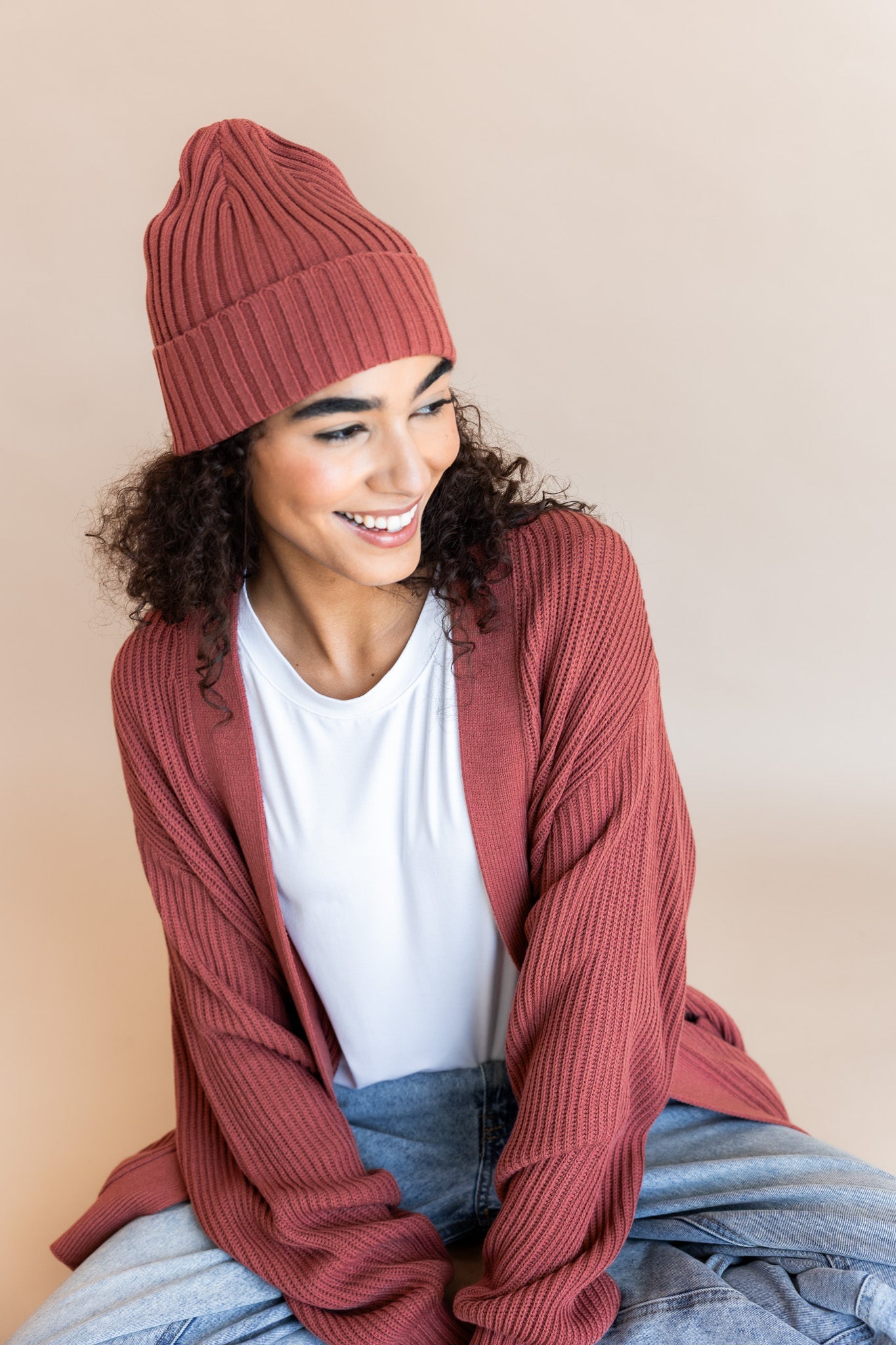 Modal wearing Chunky Knit Adult Ribbed Beanie in ember over white t-shirt and chunky knit women's cardigan in ember and blue jeans on a beige background