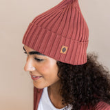 Modal wearing Chunky Knit Adult Ribbed Beanie in ember over white t-shirt and chunky knit women's cardigan in ember on a beige background