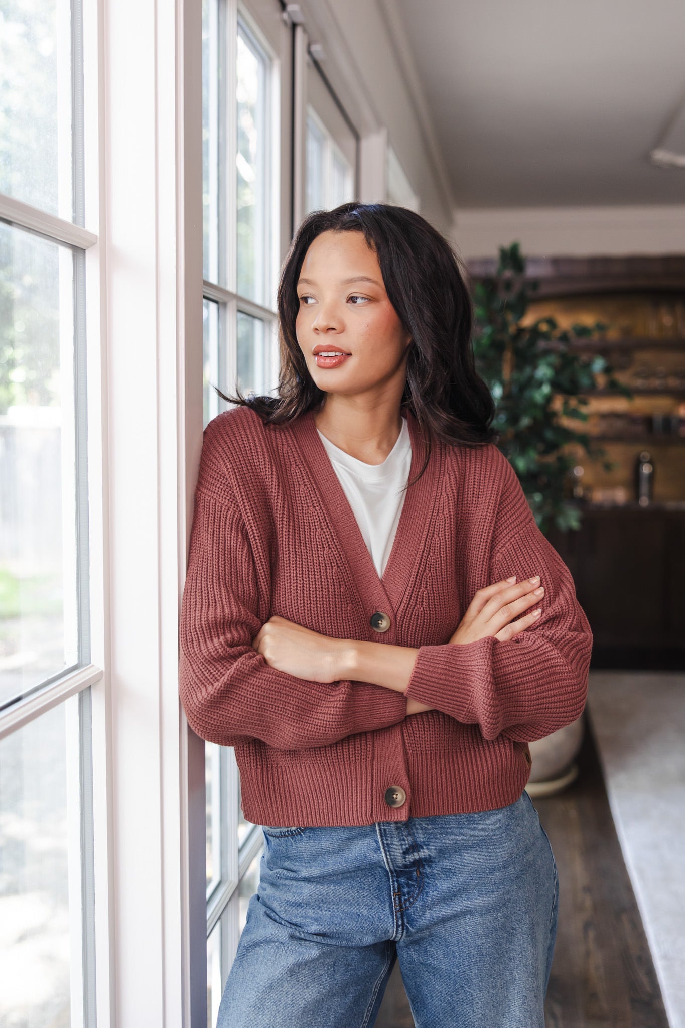 Woman leaned against the wall looking out the window wearing a cropped cardigan in ember
