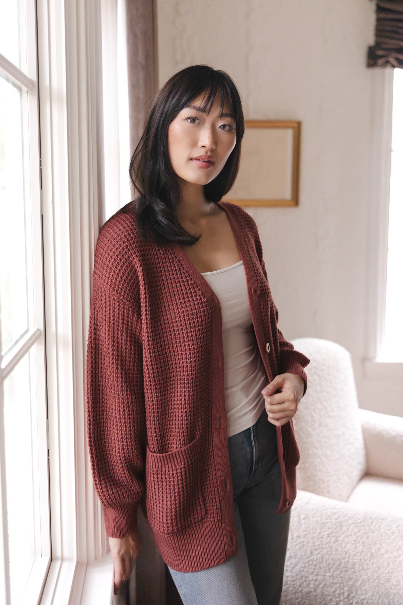 Modal standing near a window wearing Chunky Knit Women's Waffle Cardigan in Ember over white camisole and blue jeans
::: Model is 5'9" wearing size S ///