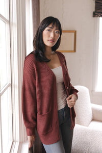 Modal standing near a window wearing Chunky Knit Women's Waffle Cardigan in Ember over white camisole and blue jeans