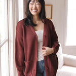 Modal smiling near a window wearing Chunky Knit Women's Waffle Cardigan in Ember over white camisole and blue jeans