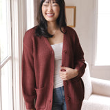 Modal smiling near a window wearing Chunky Knit Women's Waffle Cardigan in Ember over white camisole and blue jeans