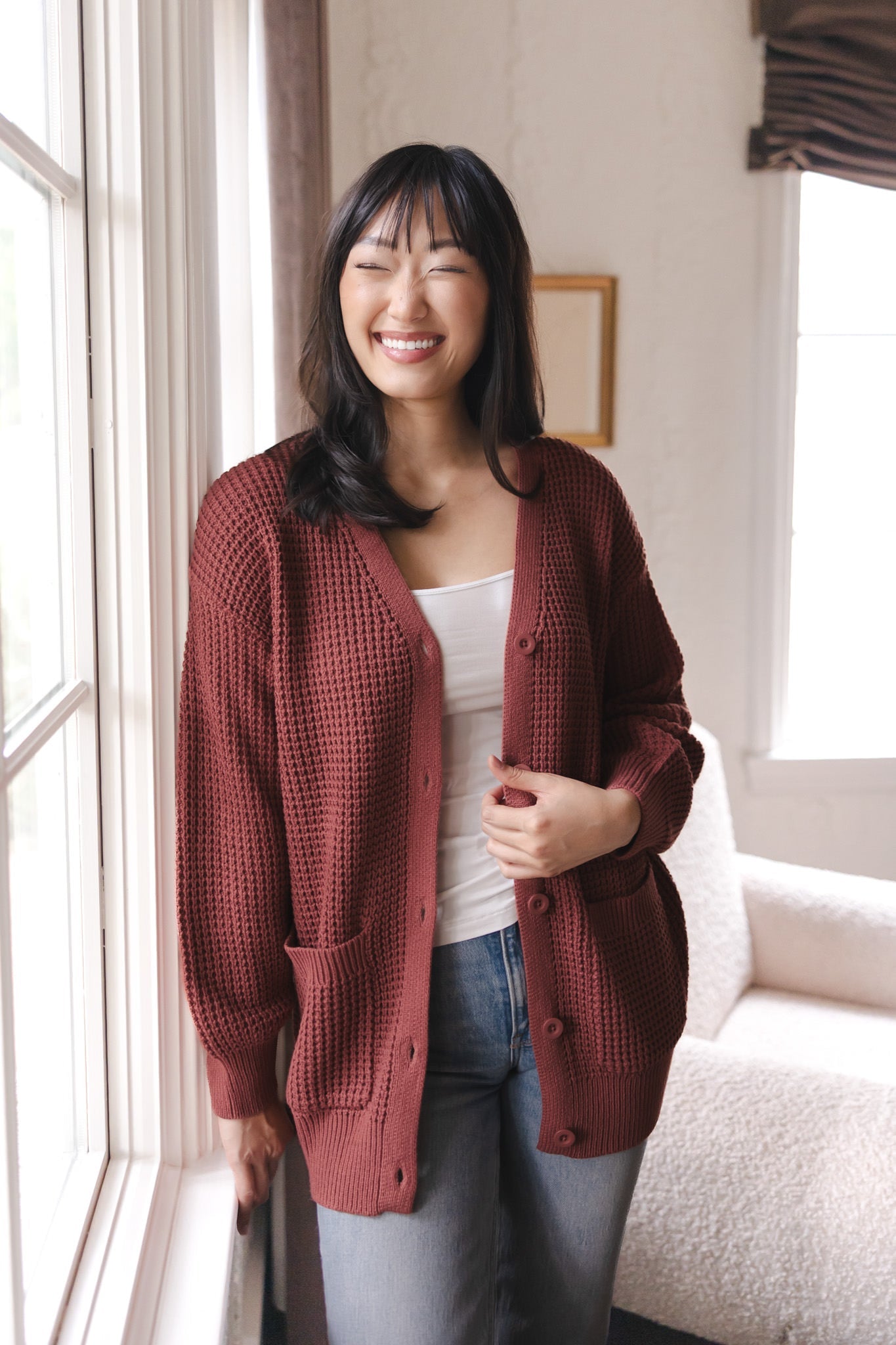 Modal smiling near a window wearing Chunky Knit Women's Waffle Cardigan in Ember over white camisole and blue jeans