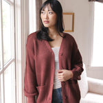 Modal standing near a window wearing Chunky Knit Women's Waffle Cardigan in Ember over white camisole and blue jeans