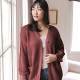 Modal standing near a window wearing Chunky Knit Women's Waffle Cardigan in Ember over white camisole and blue jeans