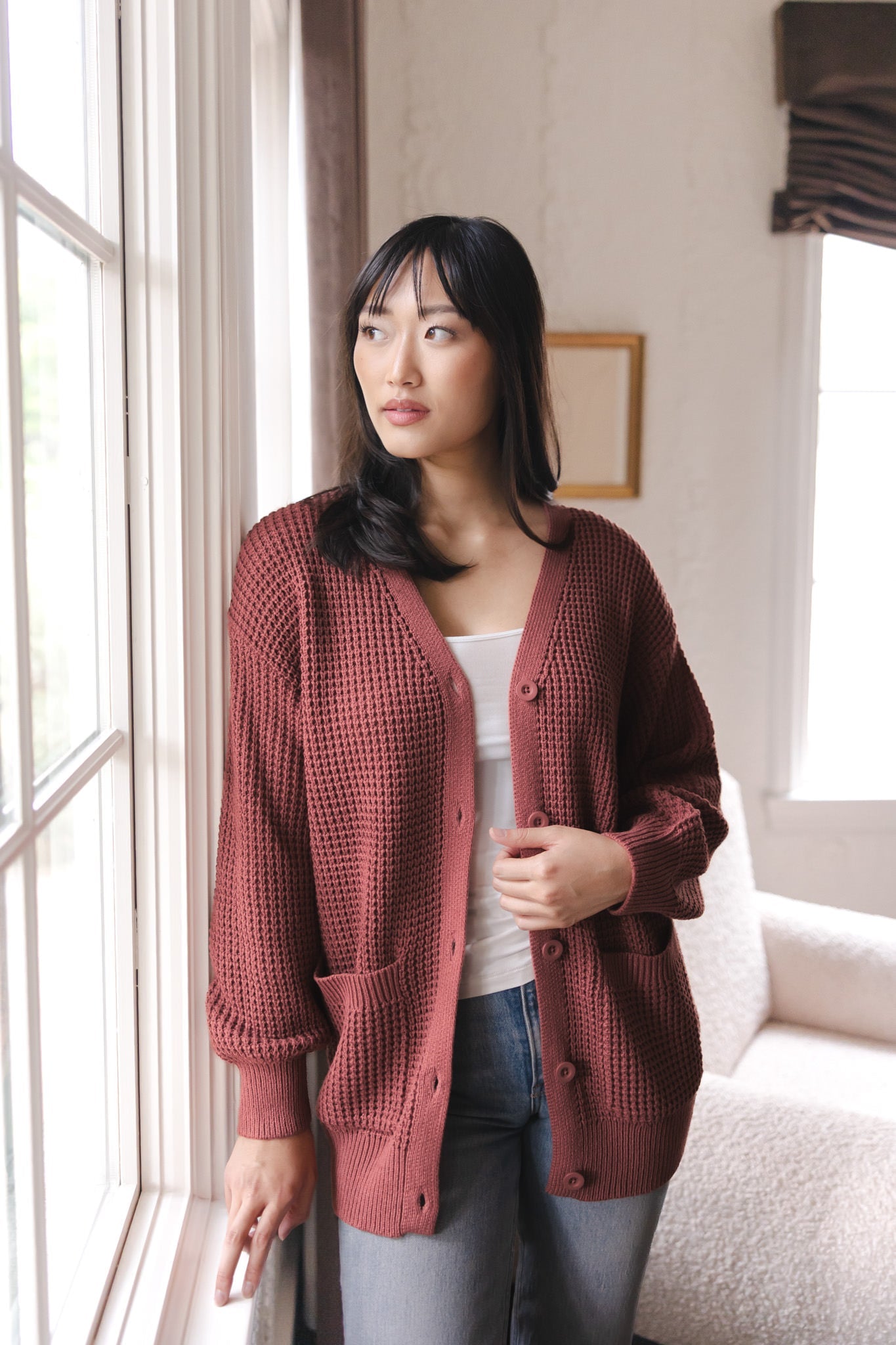 Modal standing near a window wearing Chunky Knit Women's Waffle Cardigan in Ember over white camisole and blue jeans