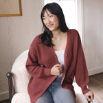 Modal sitting on a white sofa wearing Chunky Knit Women's Waffle Cardigan in Ember over white camisole and blue jeans