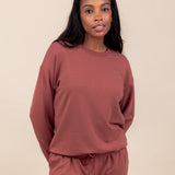 Woman wearing Women's Sweatshirt in Ember color over similar color jogger pants on a beige background