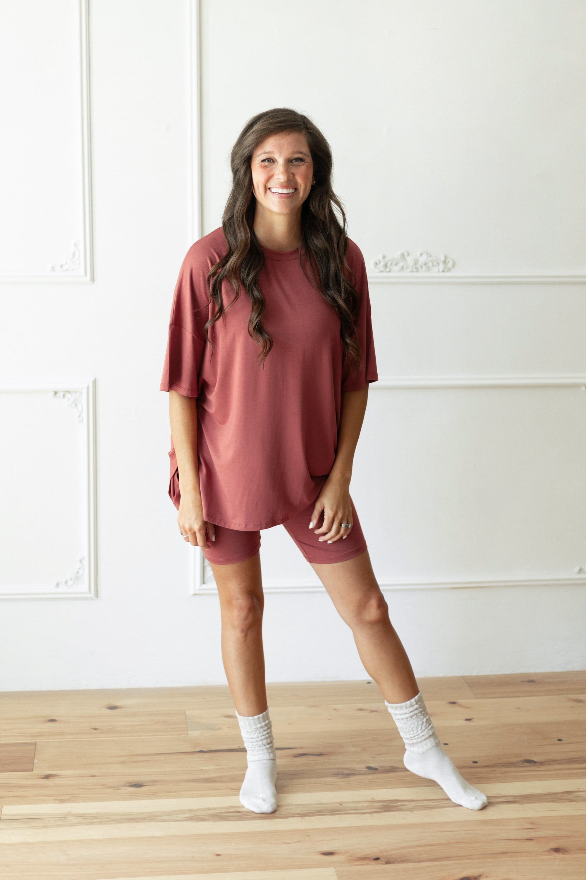 A woman modeling a dusty rose short-sleeve t-shirt and matching shorts set with white socks, standing on a light wood floor against a white paneled wall.