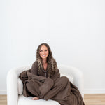 Smiling model curled up while wrapped in Model curled up on a white sofa in Kyte Quilt Blanket in Espresso 2.5 and matching pajama set
