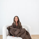 Smiling model curled up while wrapped in Model curled up on a white sofa in Kyte Quilt Blanket in Espresso 2.5 and matching pajama set