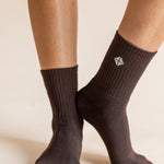 A close-up of a person's feet wearing dark brown ribbed crew socks against a warm beige background. A small white geometric logo is visible on the side of the right sock.
