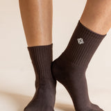 A close-up of a person's feet wearing dark brown ribbed crew socks against a warm beige background. A small white geometric logo is visible on the side of the right sock.