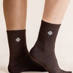 A close-up of a person's feet wearing dark brown crew socks with a small white geometric logo on the side, set against a plain light beige background.