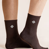 A close-up of a person's feet wearing dark brown crew socks with a small white geometric logo on the side, set against a plain light beige background.