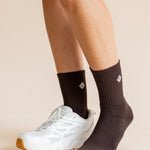 A close-up of a person's legs wearing dark brown ribbed crew socks and a white athletic sneaker against a plain beige studio background.