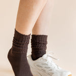 A close-up of a person's legs wearing dark brown ribbed crew socks and a white athletic sneaker against a solid beige background.