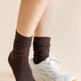 A close-up of a person's legs wearing dark brown ribbed crew socks and a white athletic sneaker against a solid beige background.