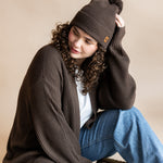 Modal wearing Chunky Knit Adult Pom in espresso over white t-shirt and chunky knit women's cardigan in espresso and blue jeans on a beige background