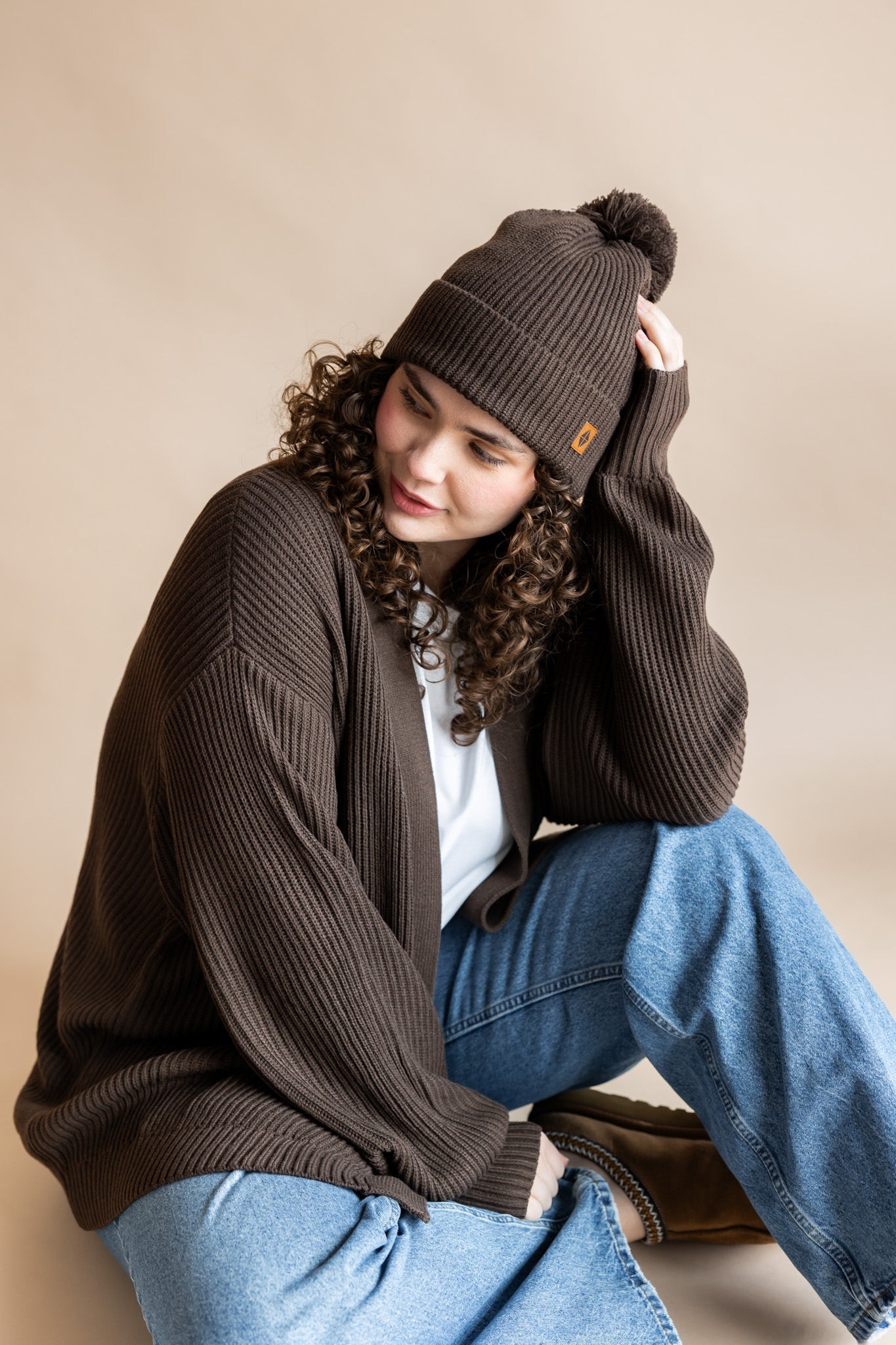 Modal wearing Chunky Knit Adult Pom in espresso over white t-shirt and chunky knit women's cardigan in espresso and blue jeans on a beige background