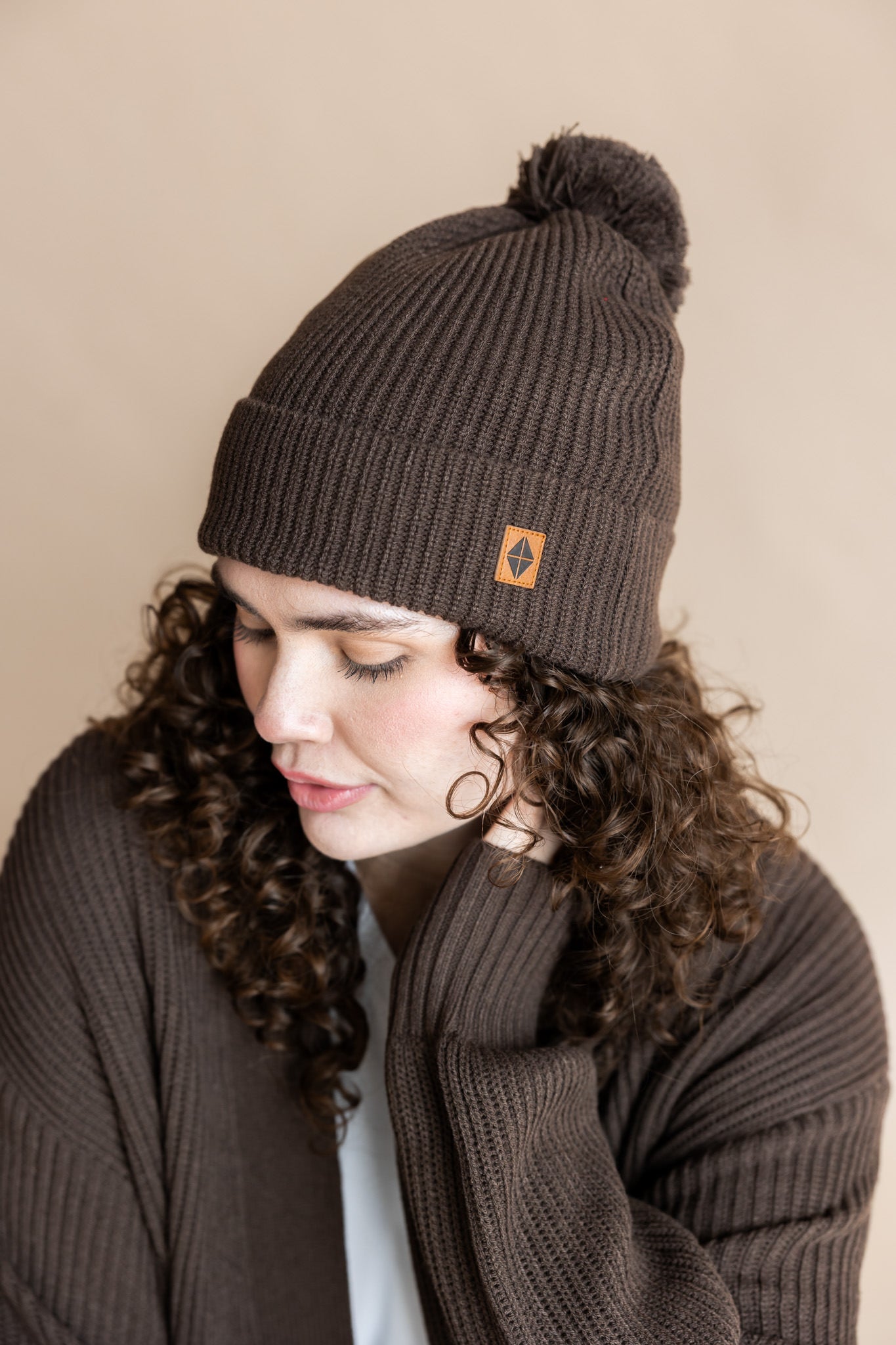 Modal wearing Chunky Knit Adult Pom in espresso over white t-shirt and chunky knit women's cardigan in espresso on a beige background